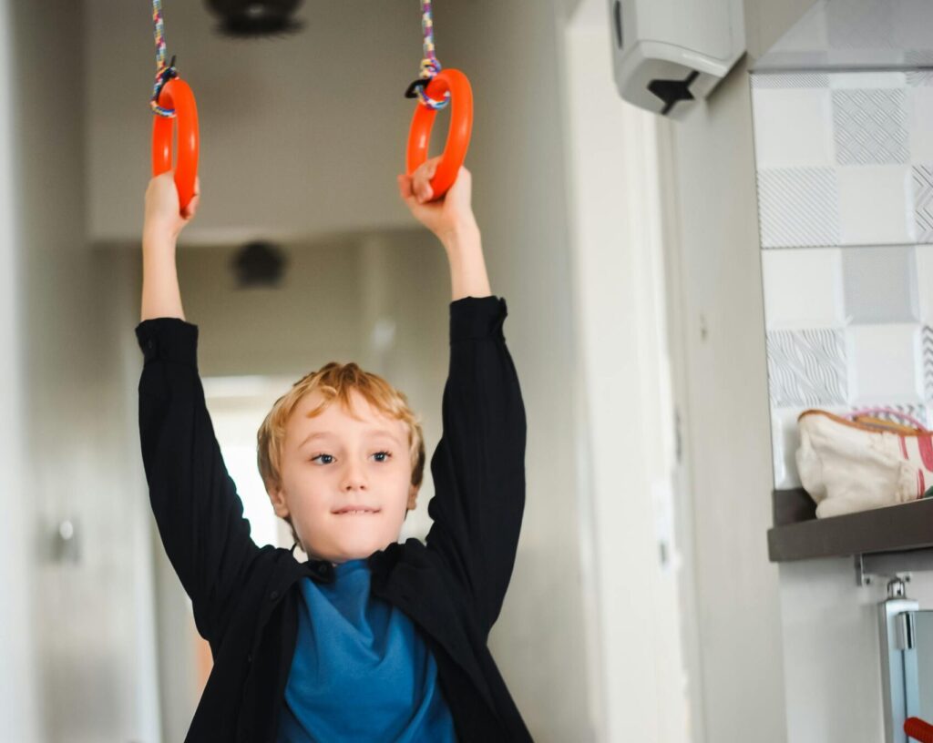 A young boy enjoying indoor play on bright orange gymnastic rings in a hallway.