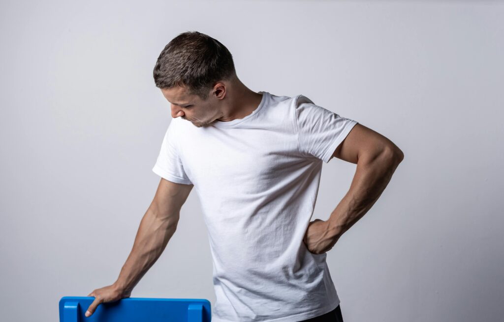 A man in a white t-shirt leans on a blue chair against a neutral backdrop.