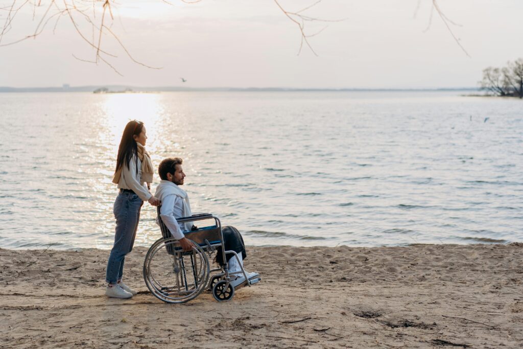 A couple enjoys a romantic moment on the beach, one in a wheelchair, with a serene seaside view.