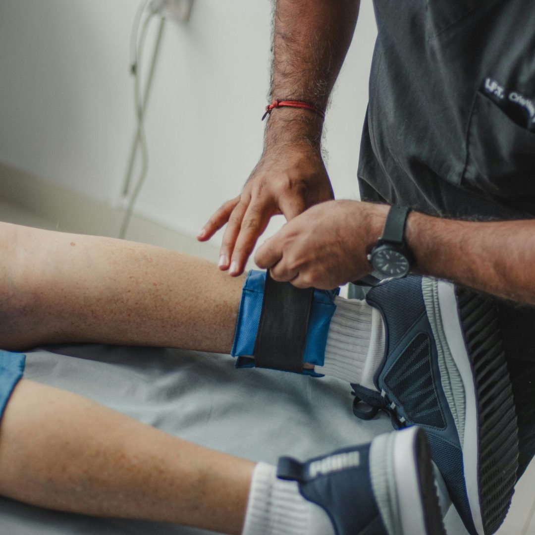 A physiotherapist adjusts a leg strap on a patient in a clinical setting.